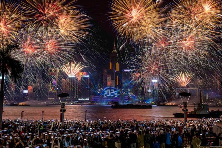 Fireworks light up a city skyline at night, marking a New Year celebration. A crowded waterfront is filled with people capturing the moment on cameras