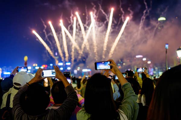 People take photos of a fireworks display in a city at night, showcasing a festive atmosphere during a travel event