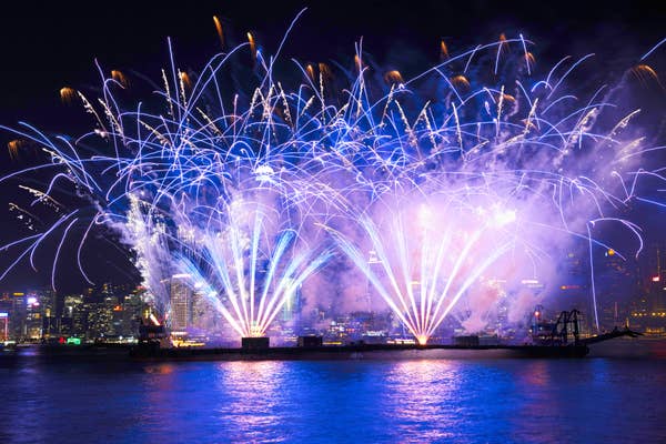 Fireworks light up the night sky over a city skyline, reflecting on the water, creating a festive atmosphere