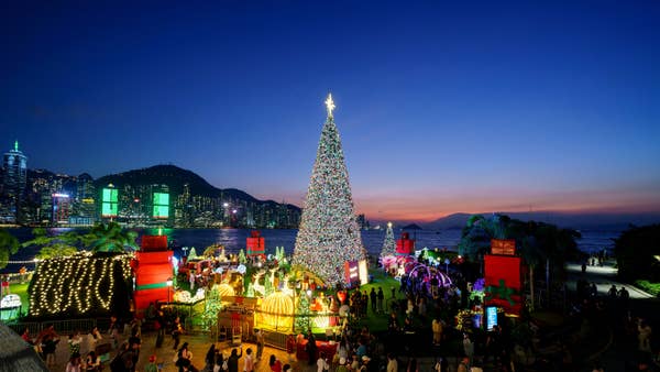 Festive crowds gather around a tall Christmas tree with city skyline in the background, under a clear evening sky