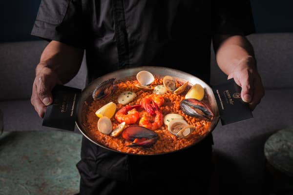 A chef presents a seafood paella, featuring mussels, clams, shrimp, and lemon slices, on a dark background