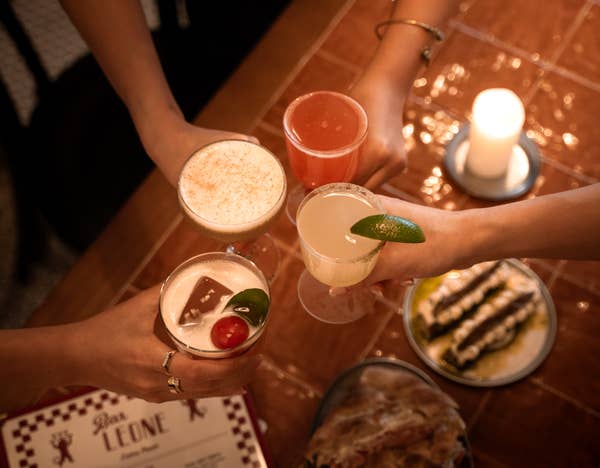 Four people toast with cocktails over a table with a candle, appetizers, and a restaurant menu