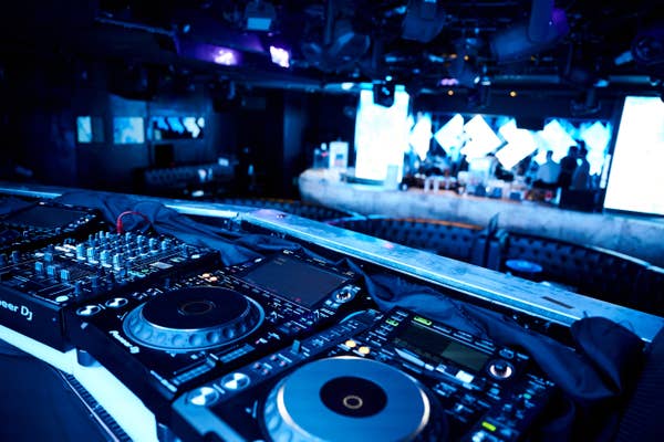 DJ booth with turntables and mixer in a vibrant, bustling nightclub setting, featuring a dance floor and stage in the background